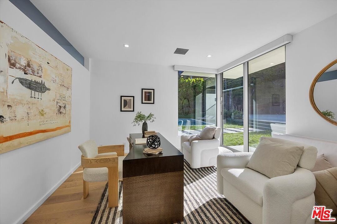 This modern home office features a sleek, dark-toned desk positioned centrally on a striped area rug, complemented by a light wood chair and two plush white armchairs. Large windows provide an expansive view of the outdoor greenery and pool area, filling the room with natural light. The space is accented by a large abstract painting on the wall and minimalist decor, creating a sophisticated and serene work environment.