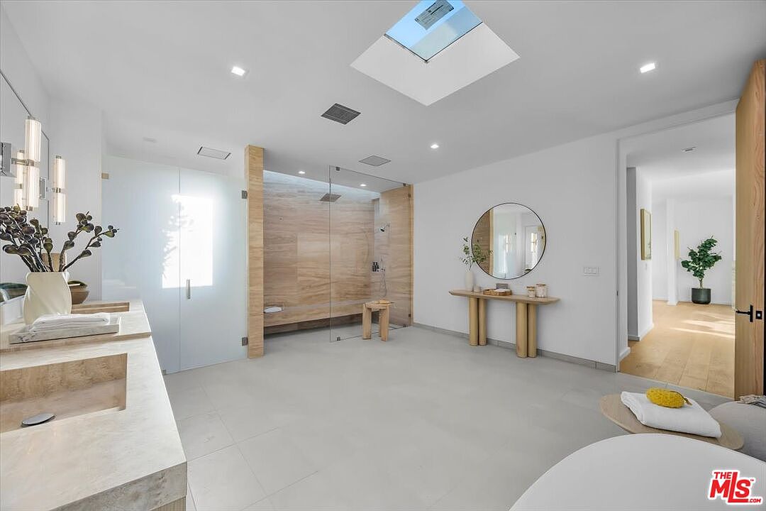 This spacious, modern primary bathroom features a large walk-in shower with light-toned stone walls and a glass enclosure. A double vanity with a stone countertop sits to the left, while a minimalist console table with a round mirror provides a decorative focal point against the white wall. The room is brightened by a skylight and recessed lighting, creating an airy and luxurious atmosphere.