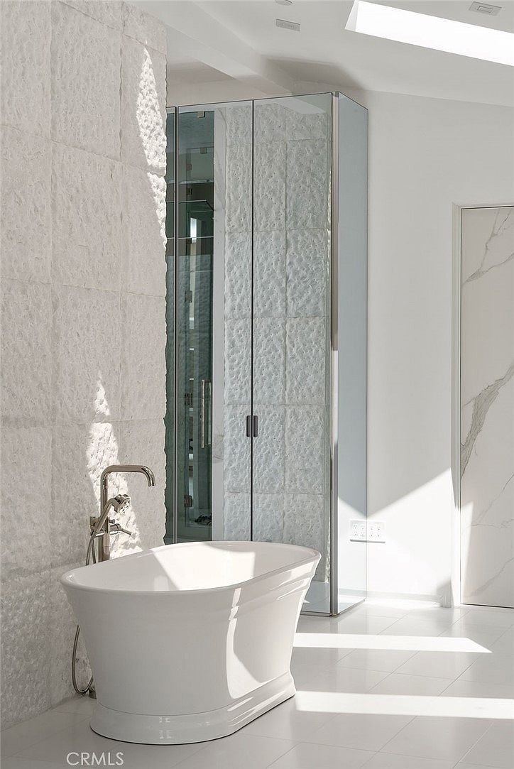 This luxurious primary bathroom features a freestanding white soaking tub positioned against a textured, light-colored stone wall. To the right, a glass-enclosed shower area is framed by mirrored panels, reflecting the bright, minimalist aesthetic of the space. The room is bathed in natural light from an overhead skylight, highlighting the clean lines and high-end finishes.