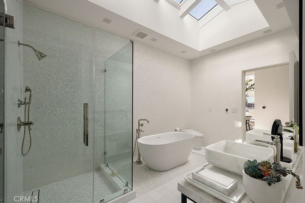 This bright and airy primary bathroom features a large glass-enclosed walk-in shower with textured wall tiles and a freestanding soaking tub positioned beneath a skylight. A modern vanity with a stone basin sink sits in the foreground, complemented by a potted succulent for a touch of greenery. The space is finished with light-toned flooring and clean, minimalist lines, creating a spa-like atmosphere.