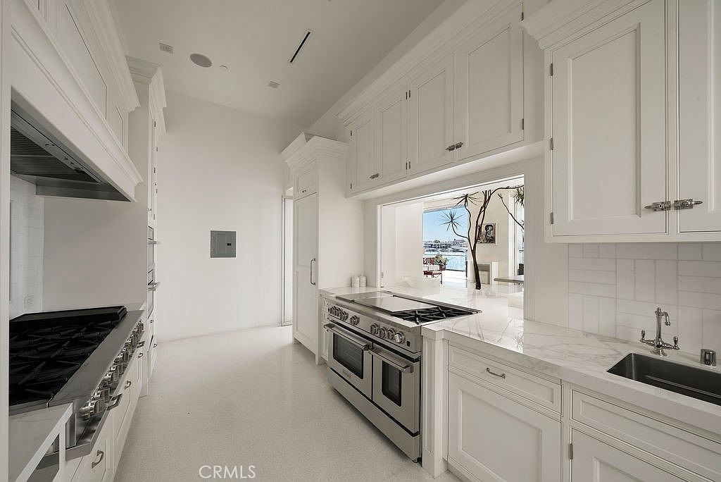 This bright, modern kitchen features crisp white cabinetry, stainless steel appliances, and light-toned stone countertops. A large pass-through window above the range provides a view of an outdoor space, while the clean lines and minimalist aesthetic create an airy, sophisticated atmosphere. The perspective highlights the galley-style layout, emphasizing the seamless integration of the cooking area with the adjacent living space.