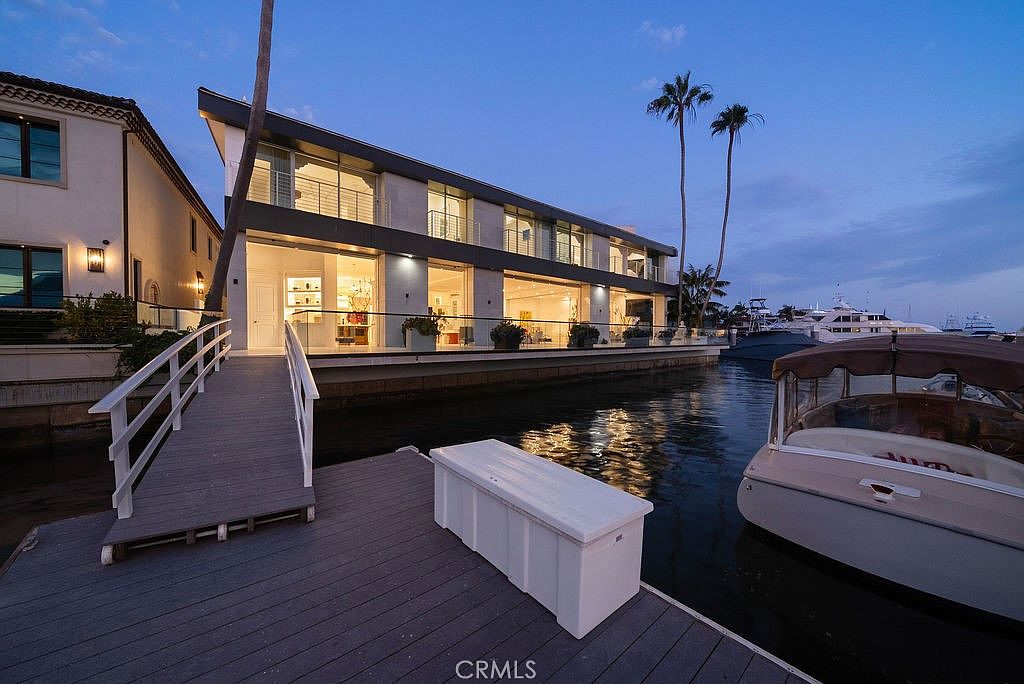 This modern waterfront residence features a sleek, contemporary design with extensive glass walls that offer a seamless connection to the canal. A private wooden dock with a storage chest extends into the water, providing direct access to a boat moored alongside the property. The twilight setting highlights the home's architectural lighting and the tranquil, upscale atmosphere of the canal-front location.