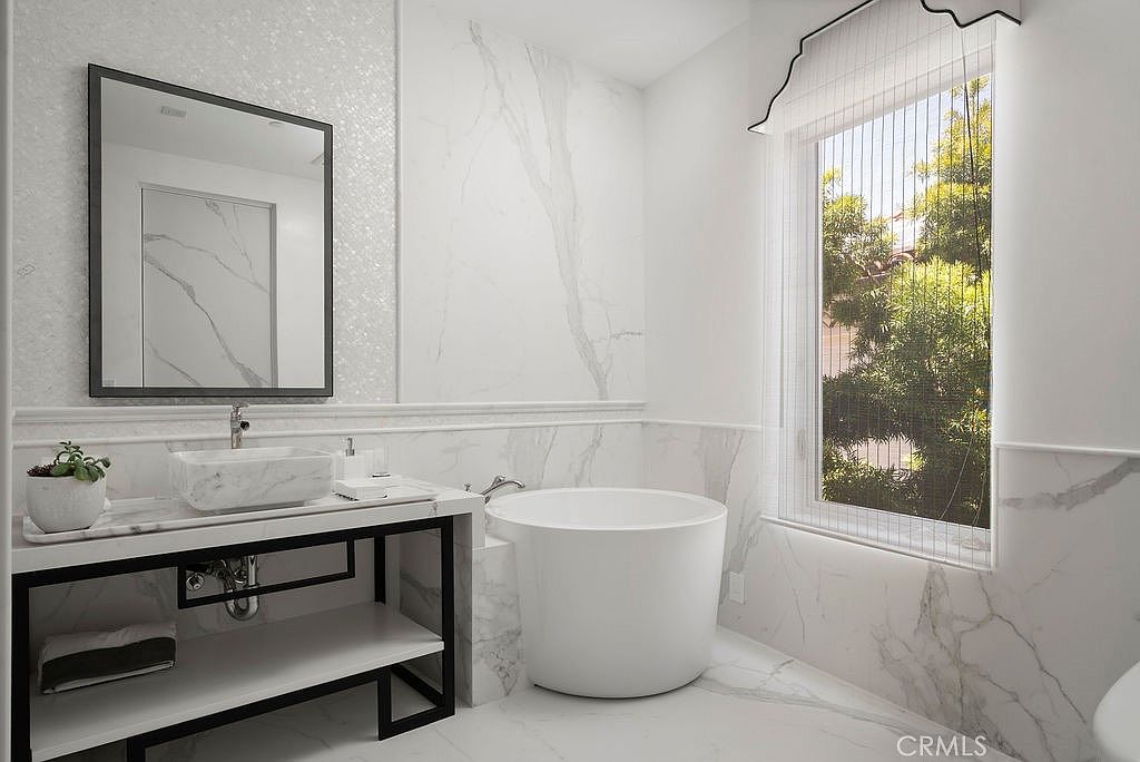 This elegant primary bathroom features a sleek, white circular soaking tub positioned next to a large window that allows for natural light. A modern vanity with a stone countertop and a rectangular vessel sink is paired with a dark metal frame, complemented by a large, framed mirror above. The walls are adorned with large-format stone tiles, creating a cohesive and luxurious aesthetic throughout the space.