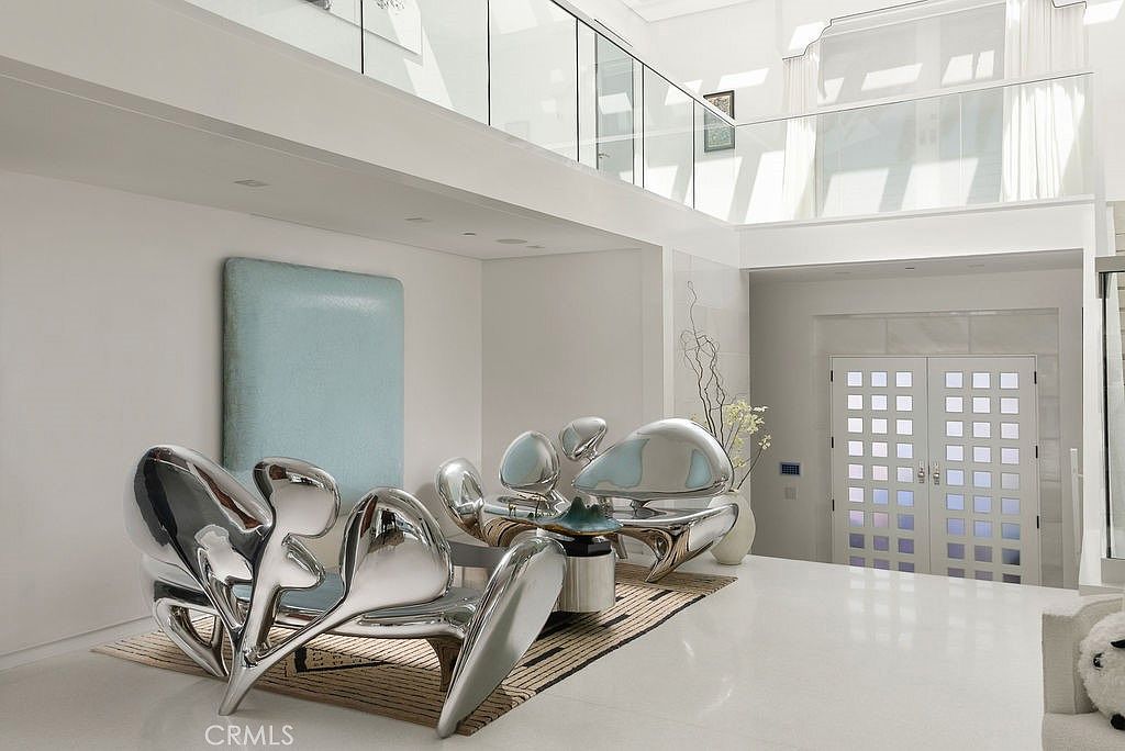 This ultra-modern, high-ceilinged living space features a striking collection of sculptural, chrome-finished furniture that serves as the room's focal point. The area is anchored by a textured area rug and a large, minimalist light blue art piece mounted on the wall. A glass-railed mezzanine overlooks the space, while a double-door entryway with a grid-patterned glass design provides a bright, architectural transition to the exterior.