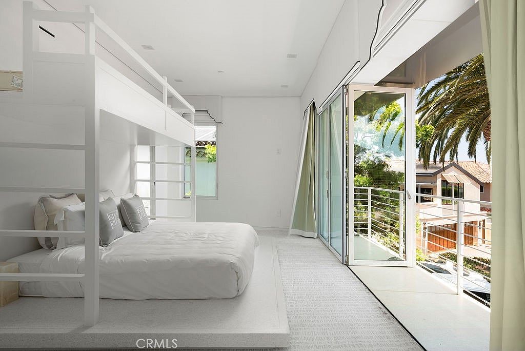 This bright, minimalist bedroom features a custom white bunk bed structure built onto a raised platform. Large, floor-to-ceiling glass doors open onto a private balcony, allowing for an abundance of natural light and a view of the surrounding neighborhood and palm trees. The room is finished in a clean, monochromatic white palette, creating an airy and modern atmosphere.