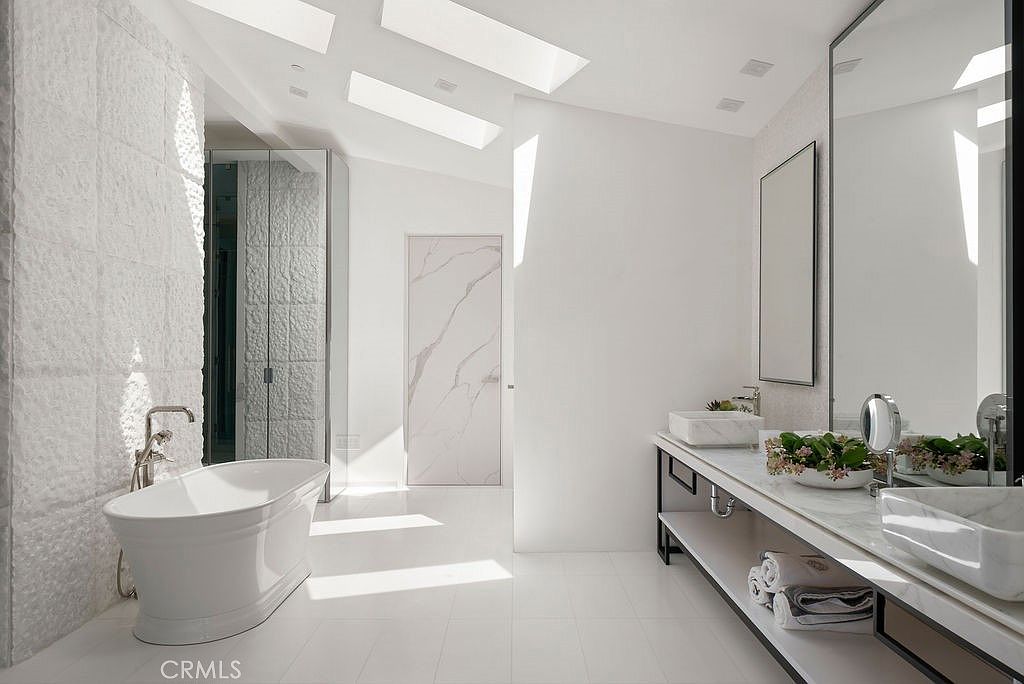 This bright, contemporary primary bathroom features a freestanding soaking tub positioned against a textured stone wall and a long vanity with dual vessel sinks. Skylights overhead flood the space with natural light, highlighting the clean lines and minimalist aesthetic. A large mirror reflects the vanity area, while a door with a subtle veined pattern adds a touch of elegance to the minimalist white interior.