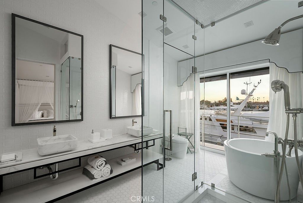This luxurious primary bathroom features a dual-vanity setup with stone-topped counters and rectangular sinks, complemented by large mirrors and a clean, white aesthetic. A glass-enclosed wet room houses a freestanding soaking tub and a shower, offering a seamless transition to a view of a yacht docked just outside the large glass sliding doors. The space is bright and modern, utilizing a neutral color palette and high-end fixtures to create a spa-like atmosphere.