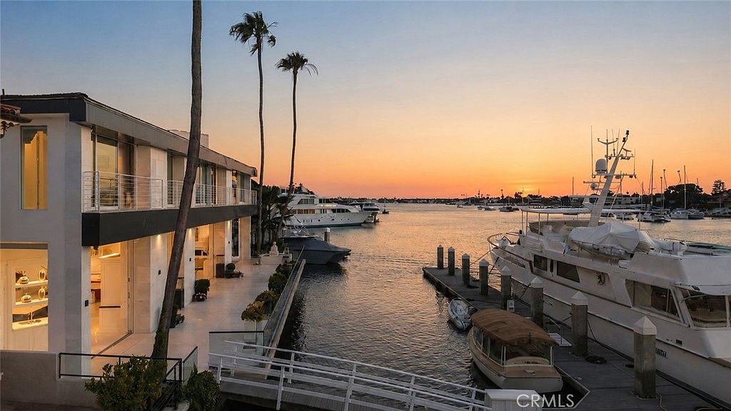 This stunning waterfront property features a modern, multi-level residence with expansive glass walls and a spacious patio overlooking a private dock. The scene is captured at sunset, casting a warm, golden glow over the calm harbor waters filled with luxury yachts and boats. The architectural design emphasizes seamless indoor-outdoor living, with a clean, contemporary aesthetic that highlights the prime location.