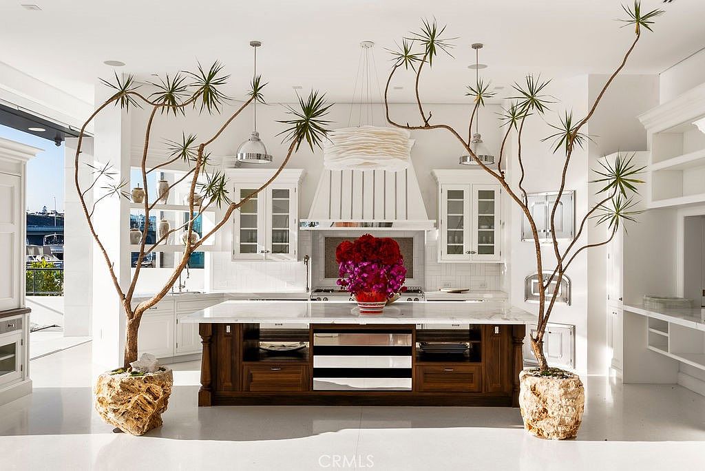 This bright, high-end kitchen features a large central island with a dark wood base and a light-colored stone countertop, topped with a vibrant floral arrangement. The space is framed by two large, sculptural potted plants that add a unique organic element to the symmetrical white cabinetry and professional-style range hood. The perspective is a centered, eye-level shot that emphasizes the clean, luxurious aesthetic and the open, airy feel of the room.