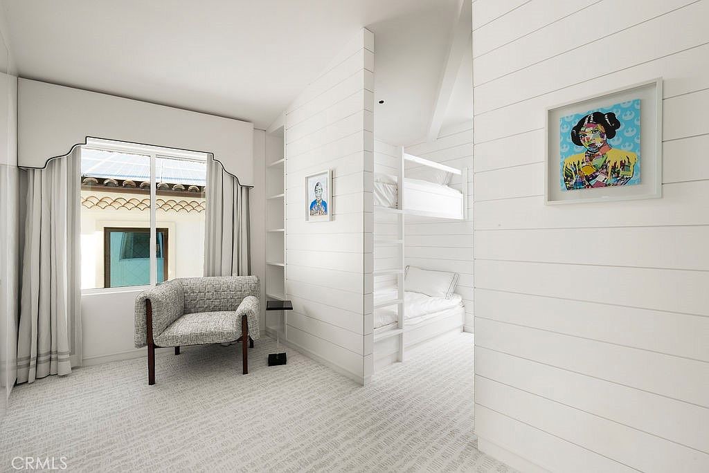 This bright, modern bedroom features white shiplap walls and a unique layout that includes a cozy reading nook with an armchair by the window and a separate alcove containing a built-in bunk bed. The room is decorated with colorful pop-art style framed prints, adding a playful touch to the clean, minimalist aesthetic. The perspective is from the main bedroom area looking toward the bunk alcove, highlighting the clever use of space and architectural detail.