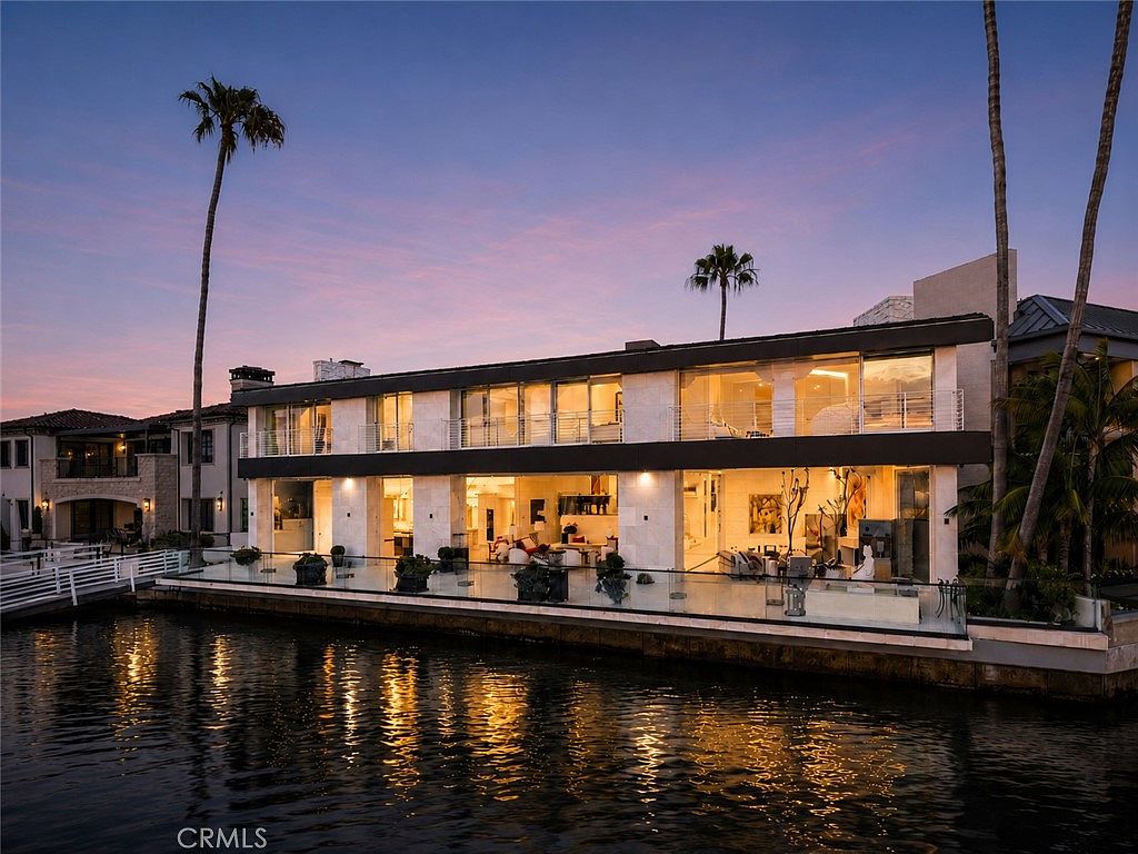 This stunning waterfront property features a contemporary architectural design with expansive glass walls that offer seamless indoor-outdoor living. The two-story structure is illuminated at dusk, highlighting its clean lines and modern aesthetic, while a glass-railed balcony overlooks the calm canal. Tall palm trees frame the scene, and the reflection of the warm interior lighting on the water creates a serene and luxurious atmosphere.