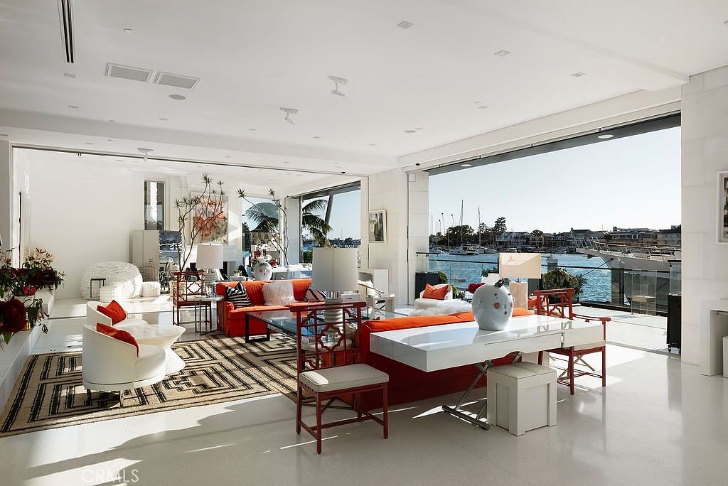 This expansive, open-concept living area features a bright, modern aesthetic with white walls and polished light-colored flooring. The space is anchored by vibrant orange sofas and a geometric patterned area rug, while floor-to-ceiling glass walls slide open to reveal a stunning waterfront view of a marina. The room is furnished with contemporary seating, decorative lamps, and a sleek white console table, creating a sophisticated and airy atmosphere perfect for luxury coastal living.