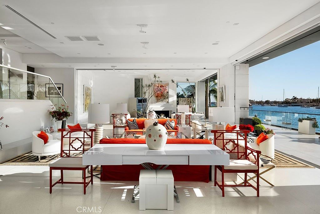 This expansive, open-concept living area features a bright, modern aesthetic with white walls and polished light-colored flooring. The space is anchored by a vibrant orange sofa and matching chairs, complemented by a central white console table topped with a decorative sculpture. Large, retractable glass walls open the room to a scenic waterfront view, seamlessly blending the indoor living space with the outdoor patio and harbor.