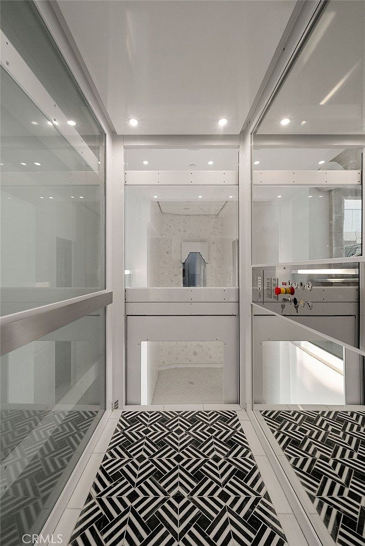 This image captures the interior of a residential elevator, featuring a modern, minimalist design with glass walls and a striking black-and-white geometric patterned tile floor. The perspective is from inside the elevator looking out toward a bright, neutral-toned space beyond the glass doors. The clean lines and reflective surfaces create a sophisticated and high-end aesthetic.
