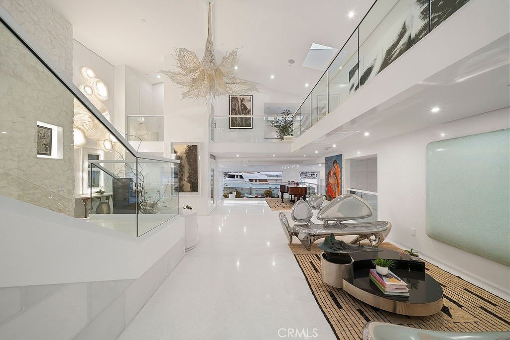 This expansive, ultra-modern living area features a high-gloss white floor and a dramatic, sculptural chandelier that serves as a focal point. The space is characterized by a double-height ceiling, a glass-railed mezzanine, and a minimalist aesthetic with avant-garde metallic furniture. Large openings at the rear provide a clear view of a yacht docked outside, emphasizing the property's waterfront location.