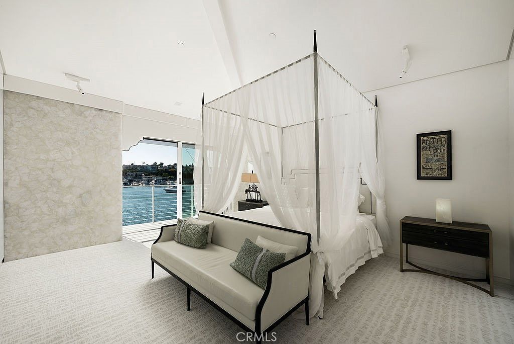 This bright and airy primary bedroom features a central canopy bed draped in sheer white fabric, positioned to face a large glass sliding door that opens to a waterfront view. A sleek, light-colored bench sits at the foot of the bed, while a minimalist console table and a framed piece of art adorn the side wall. The room is finished with a textured light-colored carpet and a distinctive, large-scale stone-textured wall panel, creating a serene and luxurious atmosphere.