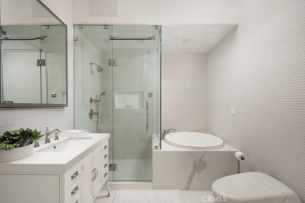 This modern primary bathroom features a white vanity with a single sink, a glass-enclosed walk-in shower, and a separate soaking tub. The walls are finished with textured horizontal-patterned tiles, and the tub surround is clad in a light-colored stone with subtle veining. A large rectangular mirror hangs above the vanity, and a white toilet is positioned to the right, creating a clean and cohesive aesthetic.