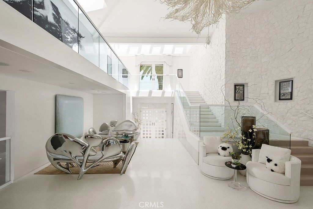 This grand, double-height entryway features a striking sculptural seating arrangement and a dramatic staircase with glass railings. The space is defined by its bright, monochromatic white palette, textured stone wall, and a large, organic-shaped chandelier suspended from the vaulted ceiling. The perspective looks directly through the foyer toward the front entrance, emphasizing the open, airy architectural design.