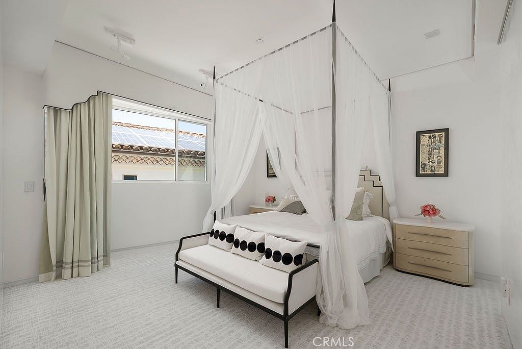 This bright and airy bedroom features a central canopy bed draped in sheer white fabric, creating a soft and elegant focal point. A modern bench with a black frame and white upholstery sits at the foot of the bed, complemented by a light-toned nightstand and a framed piece of art on the wall. The room is finished with neutral carpeting and large windows that allow natural light to fill the space, offering a view of a neighboring tiled roof.