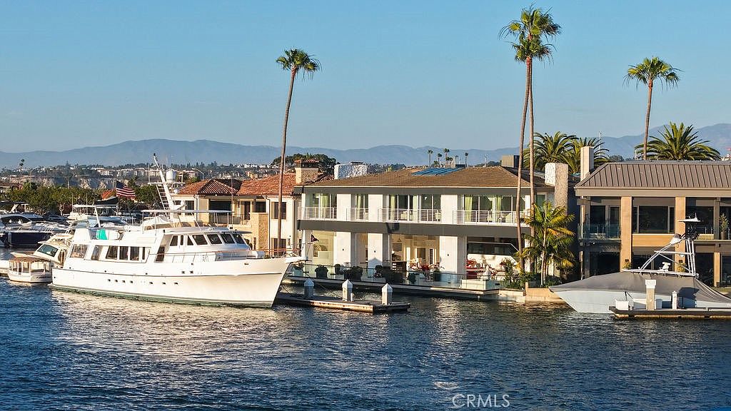 This waterfront property features a modern, multi-level residence with expansive glass walls and balconies overlooking a private dock. A large white yacht is moored alongside the home, emphasizing the luxury lifestyle associated with this harbor-front location. The scene is captured from the water, showcasing the home's architectural design and its seamless integration with the surrounding coastal environment.