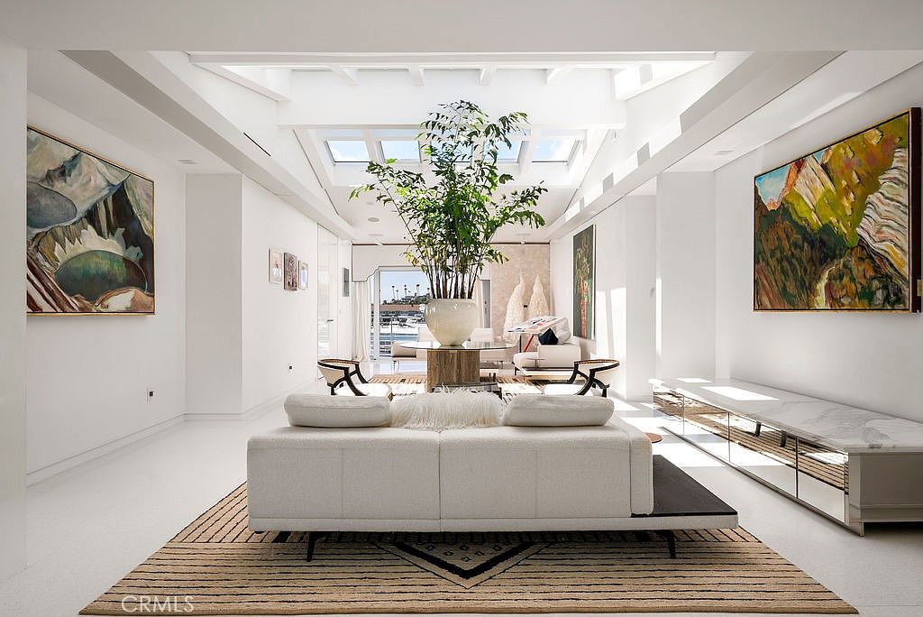 This bright and airy living room features a minimalist aesthetic with white walls, polished floors, and a large central skylight that floods the space with natural light. A contemporary white sofa sits atop a textured area rug, facing a central pedestal table topped with a large potted plant, while expansive glass doors at the rear provide a clear view of a marina. The room is accented by large, vibrant landscape paintings on either side, creating a sophisticated and serene atmosphere.