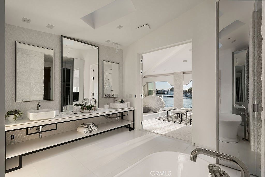 This luxurious primary bathroom features a sleek, minimalist double vanity with stone vessel sinks and a long, open-shelf base. The space is bright and airy, with a large soaking tub in the foreground and a seamless transition through an open doorway to a sunlit lounge area overlooking the water. The design emphasizes clean lines, reflective surfaces, and a sophisticated, modern aesthetic.