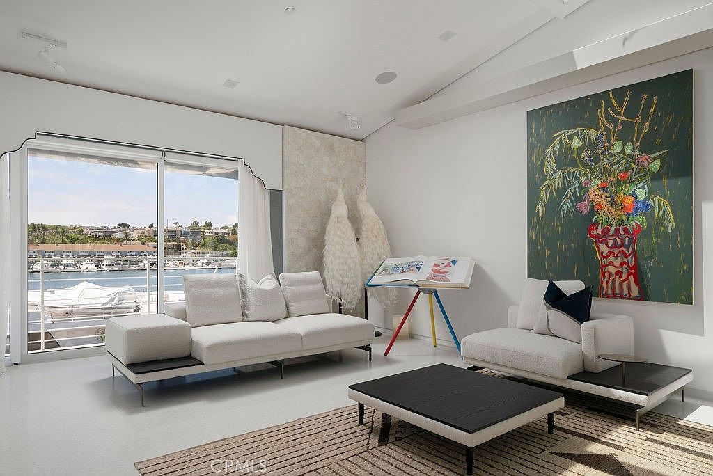 This bright, contemporary living room features a minimalist aesthetic with white walls, light-toned flooring, and a large area rug with a geometric pattern. The space is anchored by a modular white sofa and a matching armchair, complemented by a low-profile black coffee table and a unique easel-style display stand. Large sliding glass doors provide an expansive view of the harbor and boats outside, while a large, vibrant floral painting adds a bold splash of color to the room.