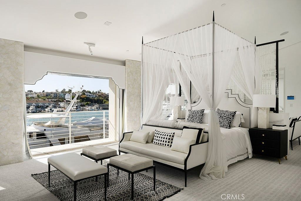 This luxurious primary bedroom features a prominent four-poster canopy bed draped in sheer white fabric, serving as the room's focal point. A sophisticated seating area with a white sofa and matching ottomans sits at the foot of the bed atop a patterned area rug, while large glass doors open directly to a balcony overlooking a marina filled with yachts. The room is decorated in a refined, neutral palette with black-and-white accents, creating an elegant and serene waterfront retreat.
