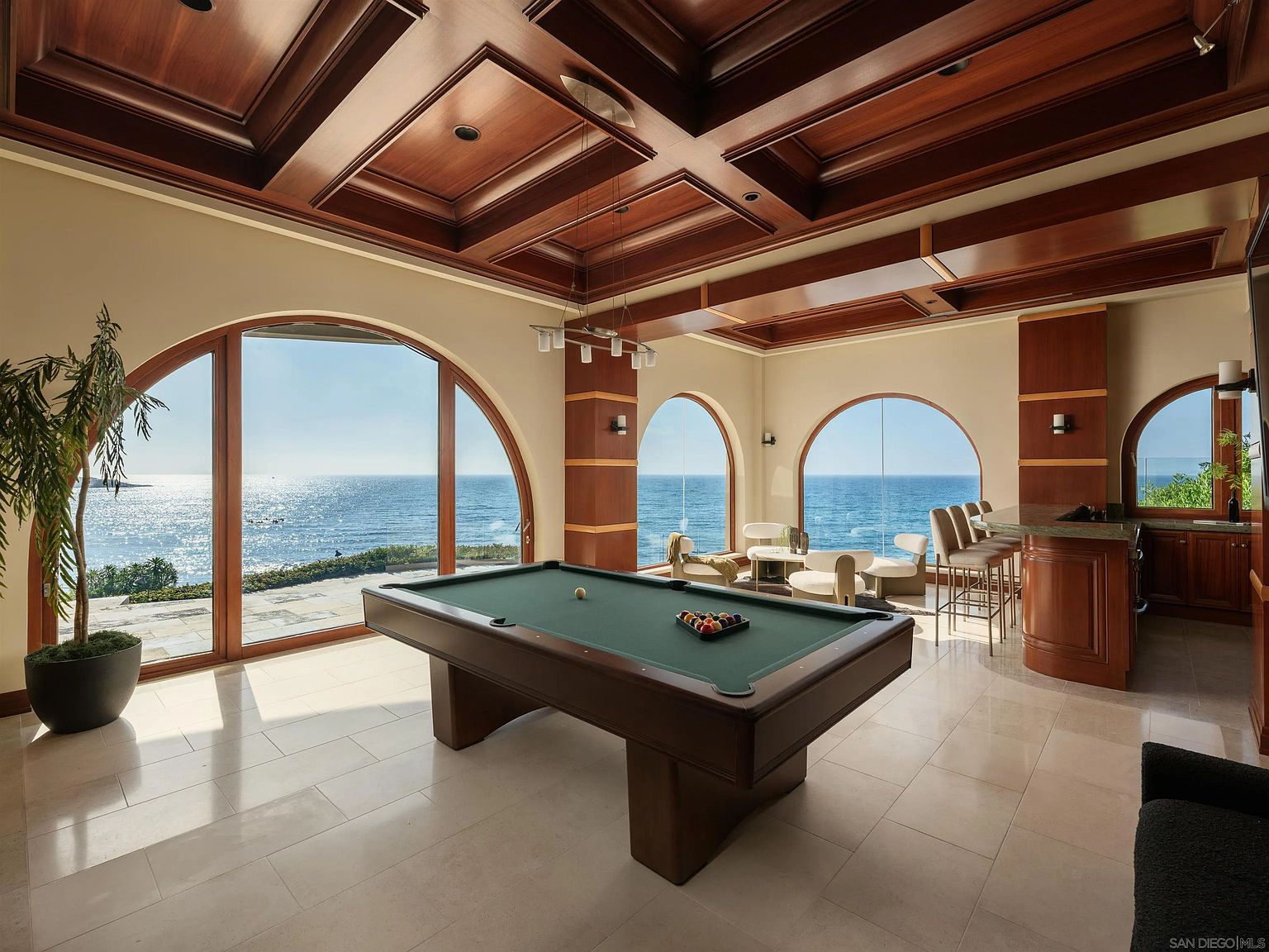 This luxurious game room features a central billiard table set against a backdrop of expansive, arched windows that provide panoramic ocean views. The room is defined by a rich, coffered wood ceiling and light-toned stone flooring, complemented by a built-in wet bar area with barstool seating. The space blends sophisticated interior design with a serene coastal atmosphere, creating an ideal environment for entertainment.