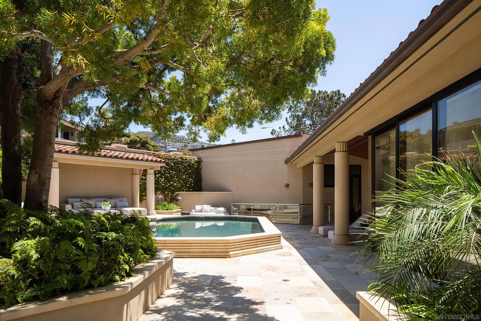 This serene outdoor living space features a geometric pool surrounded by light-toned stone paving and lush greenery. A covered patio area with comfortable seating provides a shaded retreat, while tall columns and large glass sliding doors connect the indoor and outdoor environments. The scene is framed by a mature tree, creating a peaceful and private atmosphere perfect for relaxation.