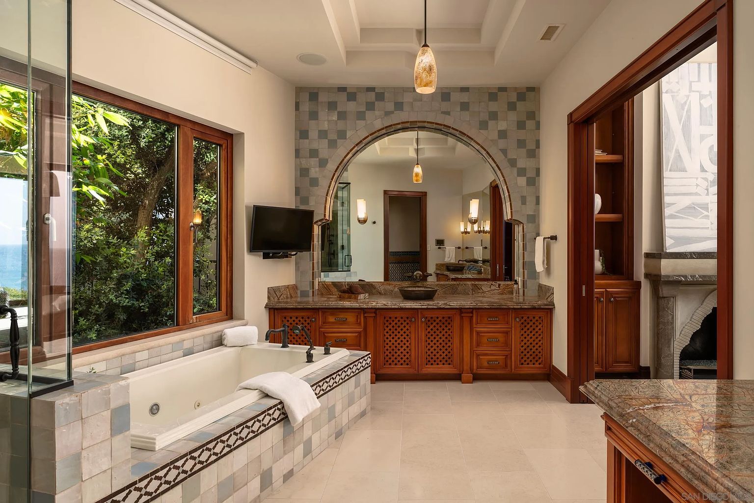 This luxurious primary bathroom features a large soaking tub set against a backdrop of patterned tile and expansive windows that offer a view of the outdoors. A central vanity with a stone countertop and a large, arched mirror serves as the focal point, complemented by warm wood cabinetry and elegant pendant lighting. The space is designed with a sophisticated, Mediterranean-inspired aesthetic, creating a serene and spa-like atmosphere.