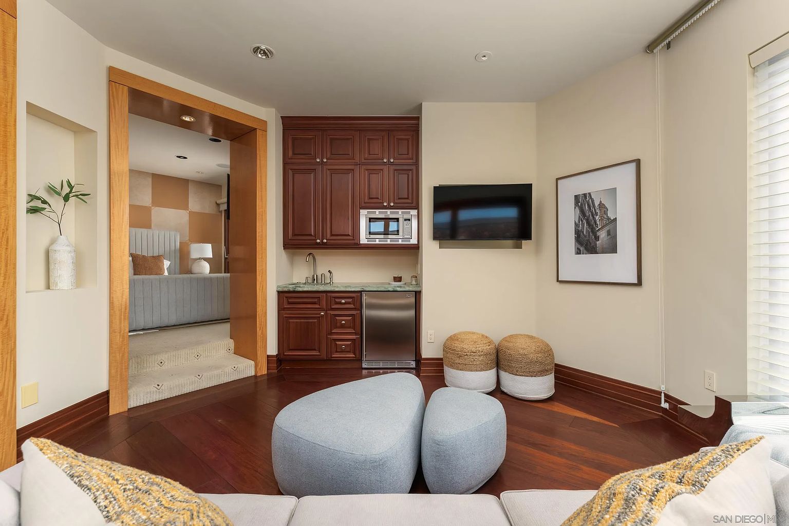 This interior space features a wet bar area with dark wood cabinetry, a small sink, and a built-in microwave, set against a neutral wall. A wall-mounted television is positioned next to a framed piece of art, while two woven ottomans and two soft, pebble-shaped ottomans provide casual seating on the dark wood flooring. A wide, wood-trimmed doorway leads up a short set of carpeted stairs into an adjacent bedroom, creating an open and connected floor plan.