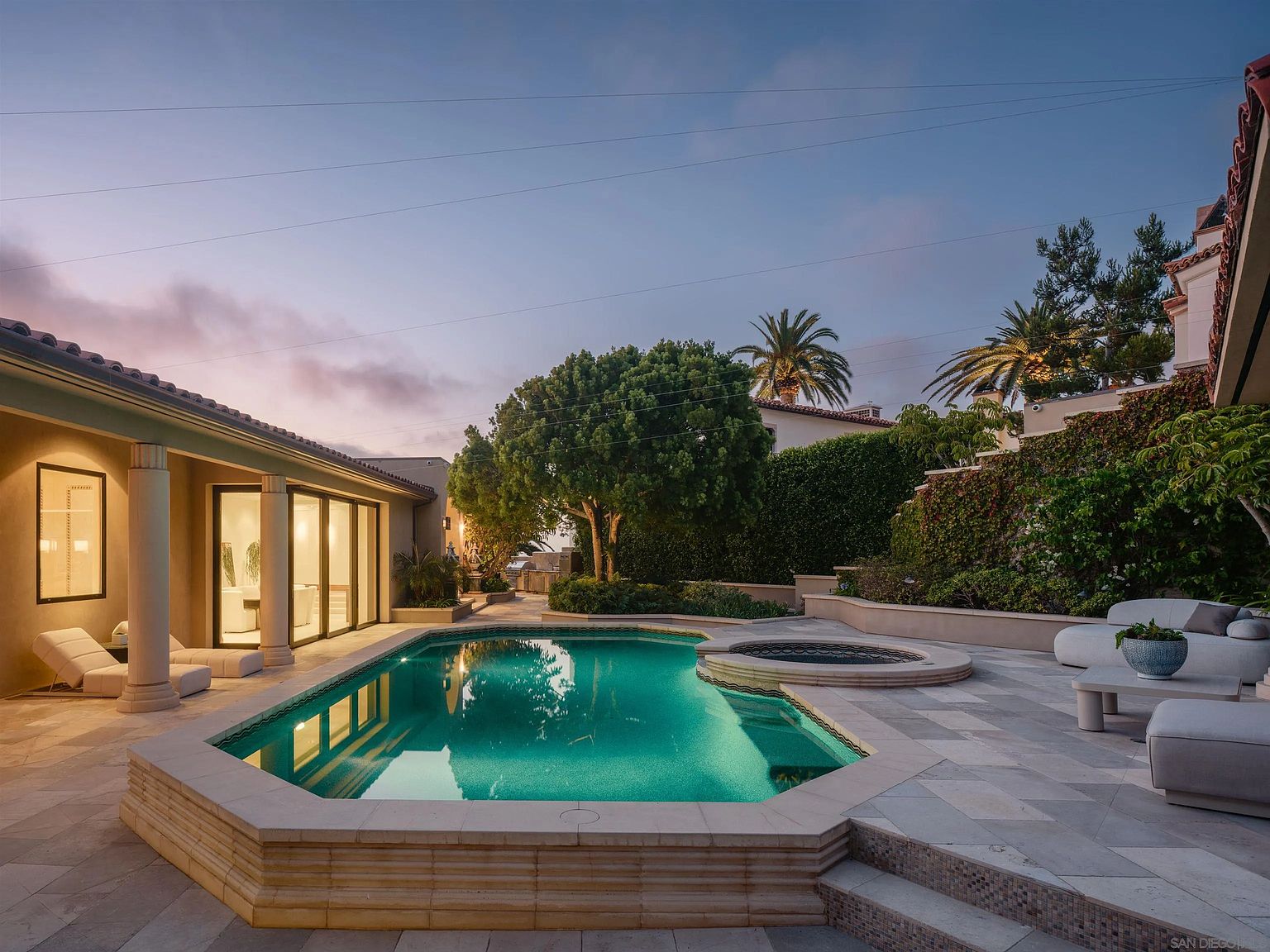 This elegant backyard features a custom-shaped swimming pool with an integrated circular spa, surrounded by light-toned stone decking. The space is framed by a covered patio with columns, lush greenery, and tall hedges, creating a private and serene outdoor retreat. The scene is captured at dusk, with warm ambient lighting highlighting the pool's turquoise water and the surrounding architectural details.