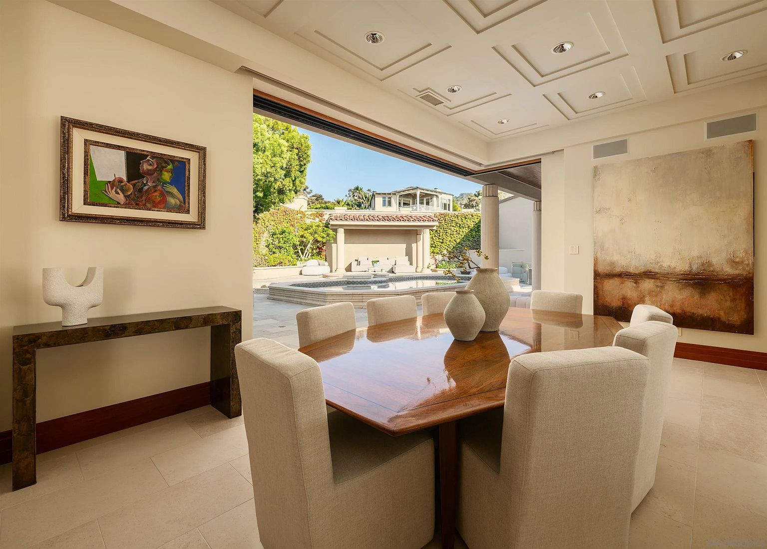 This elegant dining room features a polished wood table surrounded by six upholstered chairs, creating a sophisticated space for formal meals. The room opens directly to an outdoor patio and pool area through a wide, seamless transition, blending indoor and outdoor living. Neutral walls, a coffered ceiling, and curated artwork contribute to a refined, high-end aesthetic.
