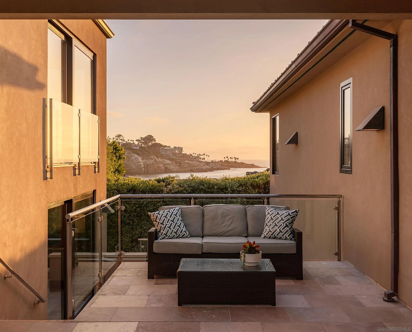 This elevated balcony offers a serene outdoor living space with a comfortable sofa and a central coffee table, framed by modern glass railings. The perspective looks out toward a picturesque coastal cove at sunset, with the warm light reflecting off the water and surrounding cliffs. The neutral-toned stone flooring and clean architectural lines create a sophisticated, tranquil atmosphere perfect for relaxation.