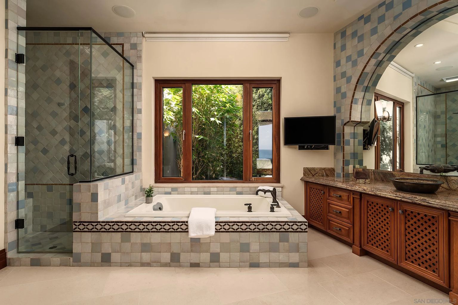 This primary bathroom features a large soaking tub set within a tiled surround, positioned beneath a set of windows that look out onto lush greenery. To the left is a glass-enclosed shower with patterned tile work, while a wooden vanity with a stone countertop and a vessel sink sits to the right. The room is accented by an arched mirror and decorative tile details, creating a warm and inviting atmosphere.