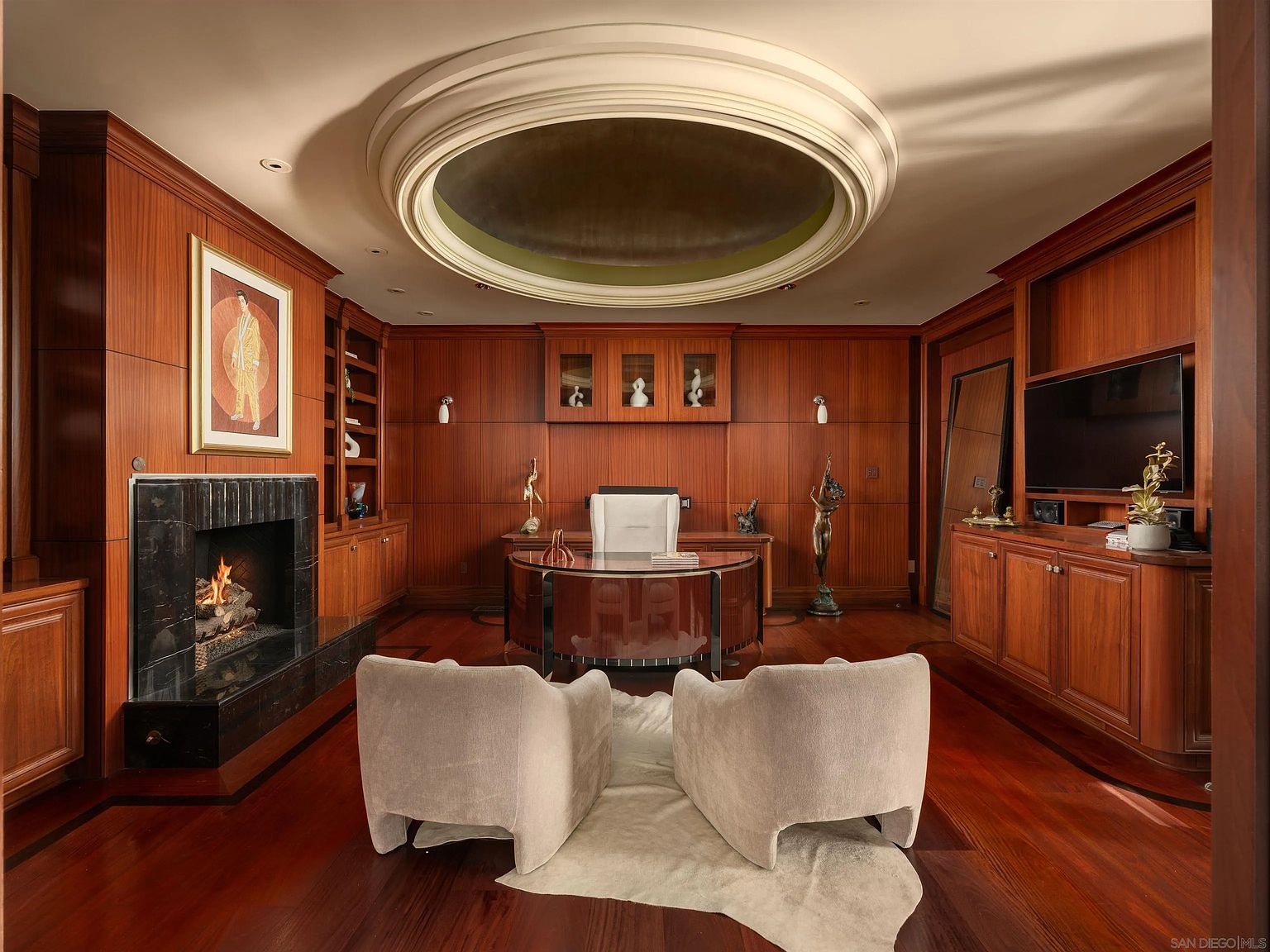 This sophisticated home office features rich wood-paneled walls, a central desk, and two plush armchairs arranged on a light-colored rug. A prominent circular recessed ceiling detail adds architectural interest, while a fireplace with a dark stone surround provides a focal point on the left. The room is elegantly furnished with built-in cabinetry, display shelving, and decorative sculptures, creating a professional yet luxurious atmosphere.