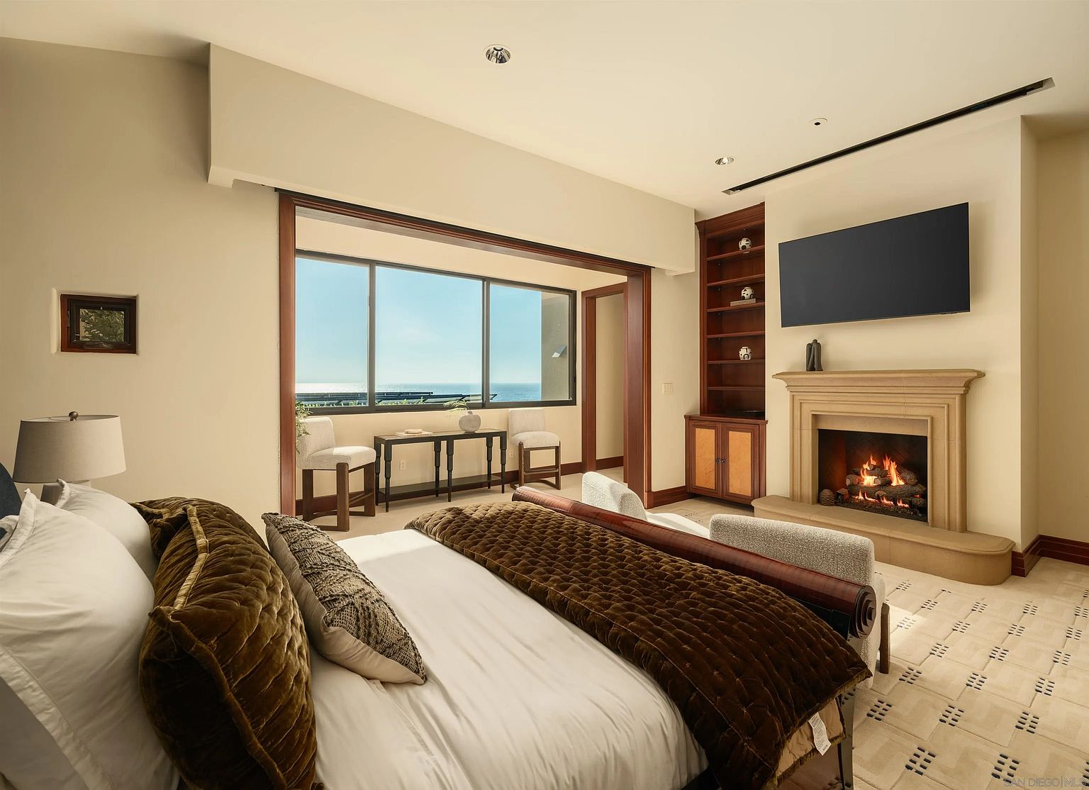 This primary bedroom features a plush bed with a dark velvet quilt, positioned to face a stone fireplace with a mounted television above. To the left, a large window wall provides an expansive view of the ocean, complemented by a console table and two chairs. The room is finished with neutral walls, a patterned carpet, and built-in wooden shelving, creating a sophisticated and tranquil atmosphere.