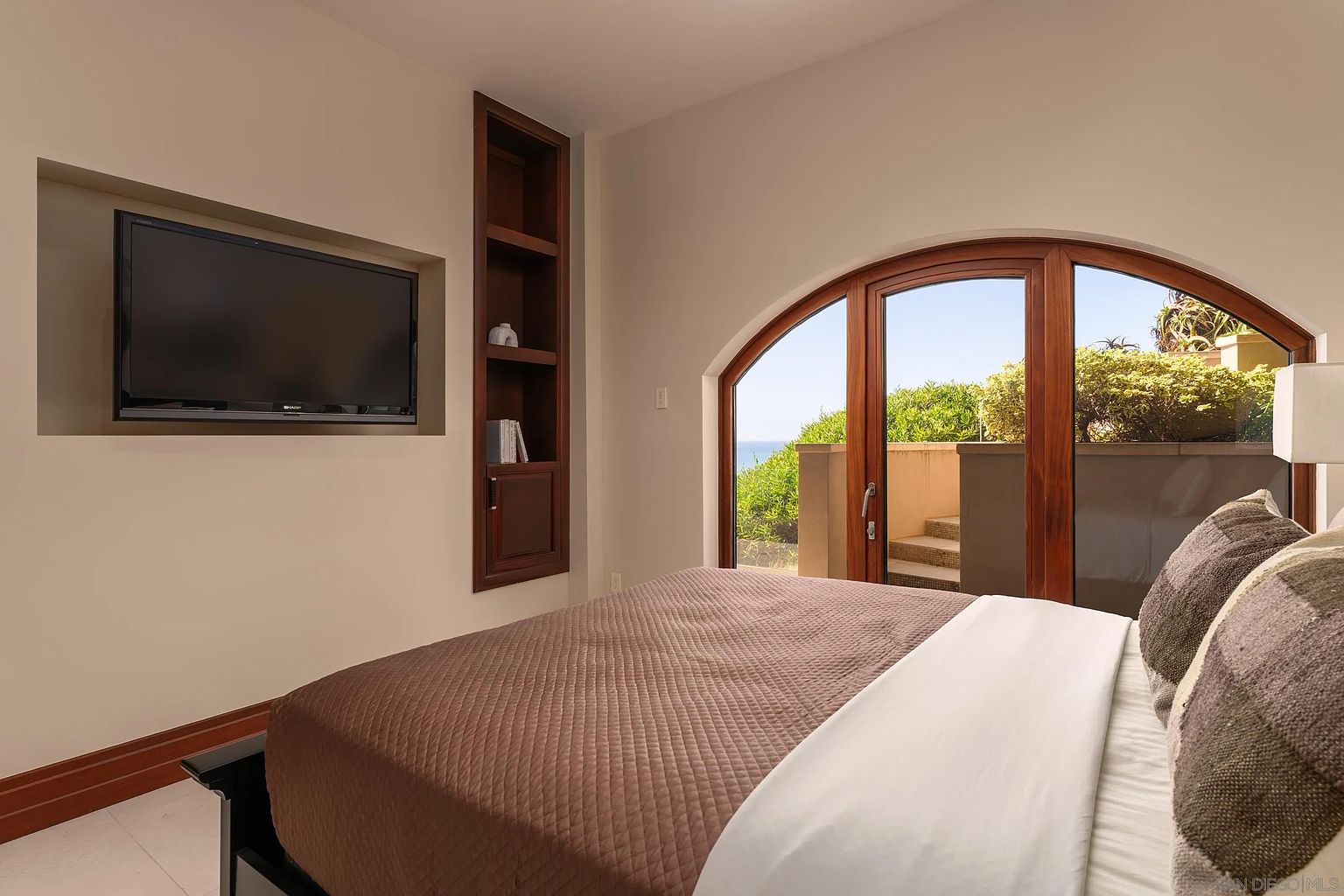 This bedroom features a neutral color palette and a large, arched wooden door that opens to an outdoor patio area with ocean views. A recessed wall niche houses a flat-screen television, complemented by a built-in wooden shelving unit for storage or display. The room is anchored by a bed with a brown quilted coverlet and patterned pillows, creating a serene and comfortable atmosphere.