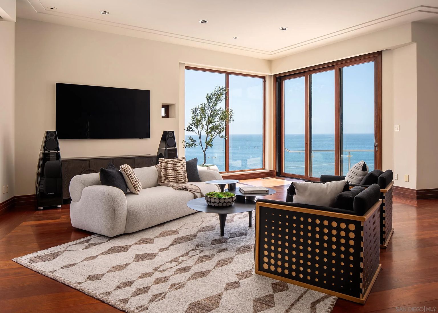 This sophisticated living room features a contemporary cream-colored sofa and two modern black-framed armchairs arranged around a tiered coffee table on a patterned area rug. The space is anchored by rich wood flooring and large windows that provide an expansive, unobstructed view of the ocean. A large wall-mounted television and sleek audio speakers complete the entertainment setup, creating a refined and tranquil atmosphere.