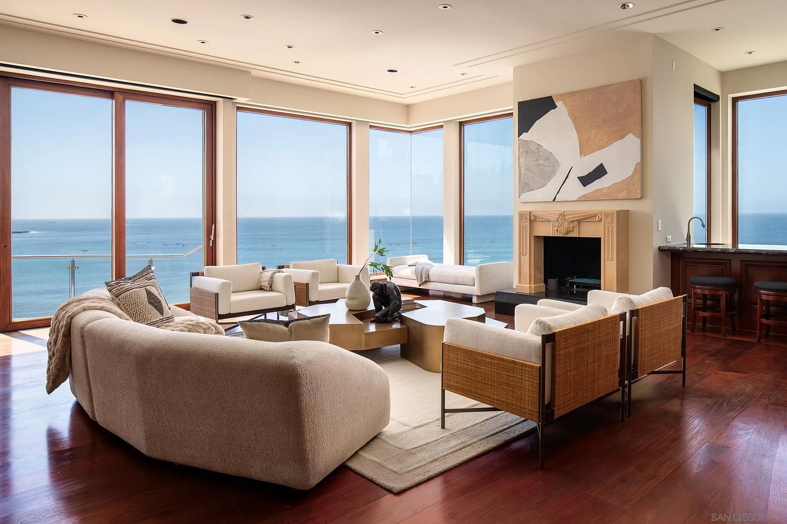 This expansive living room features a sophisticated, neutral color palette and a curved sofa that anchors the seating area. Large windows provide panoramic ocean views, while a central fireplace with a decorative mantel serves as a focal point. The space is finished with rich wood flooring and a contemporary art piece, creating an atmosphere of refined coastal luxury.