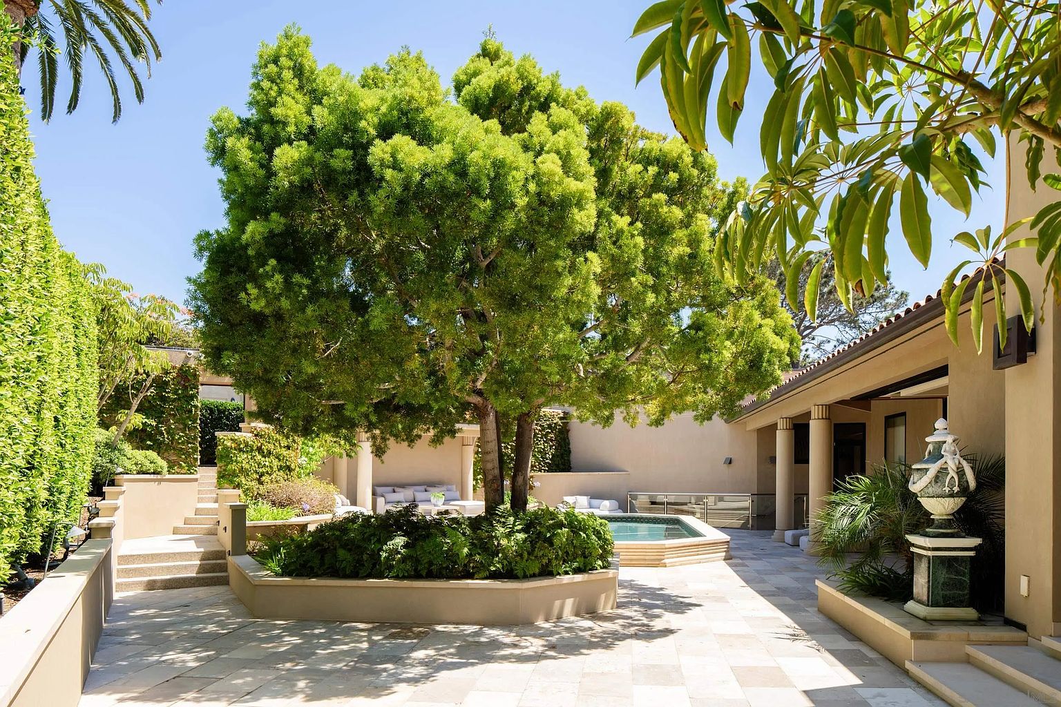This serene outdoor courtyard features a central raised planter bed housing a large, mature tree, surrounded by a stone-paved patio area. To the right, a covered loggia with columns provides a shaded seating space, while a small, built-in spa sits adjacent to the tree. The scene is framed by lush greenery and stone steps, creating a private, resort-like atmosphere perfect for relaxation.