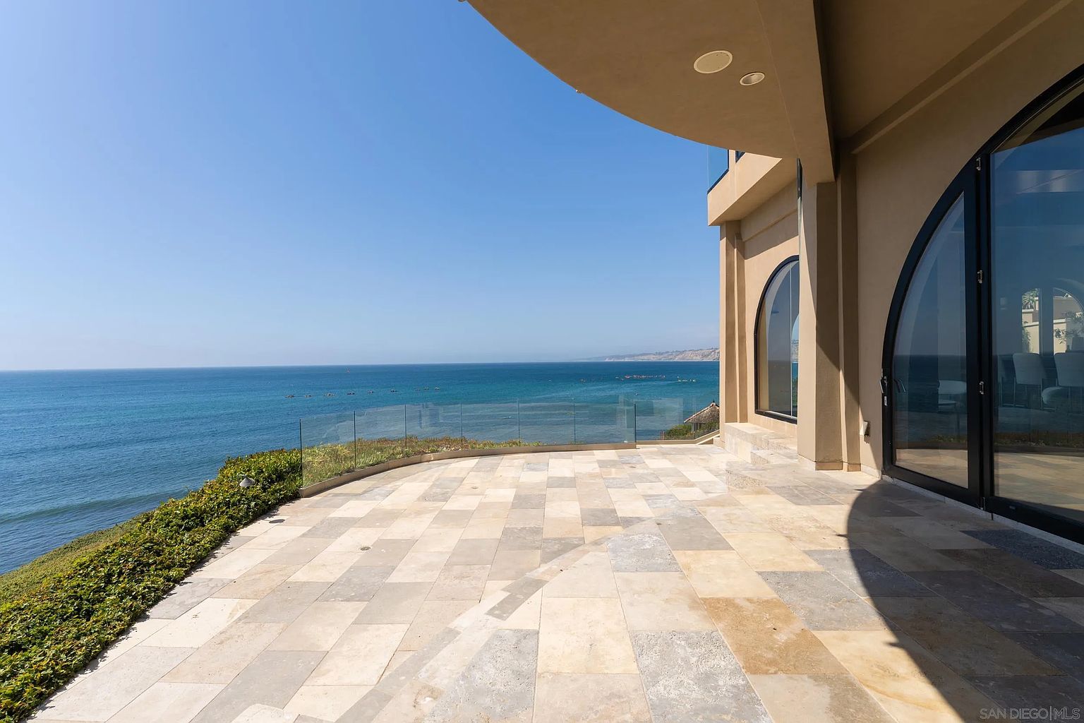 This expansive stone-tiled terrace offers a panoramic, unobstructed view of the ocean, framed by a sleek glass railing. The architectural design features a curved overhang and large, arched glass doors that seamlessly connect the indoor living space to the outdoor environment. The perspective captures the serene coastal setting, emphasizing the property's prime location and luxurious outdoor living potential.