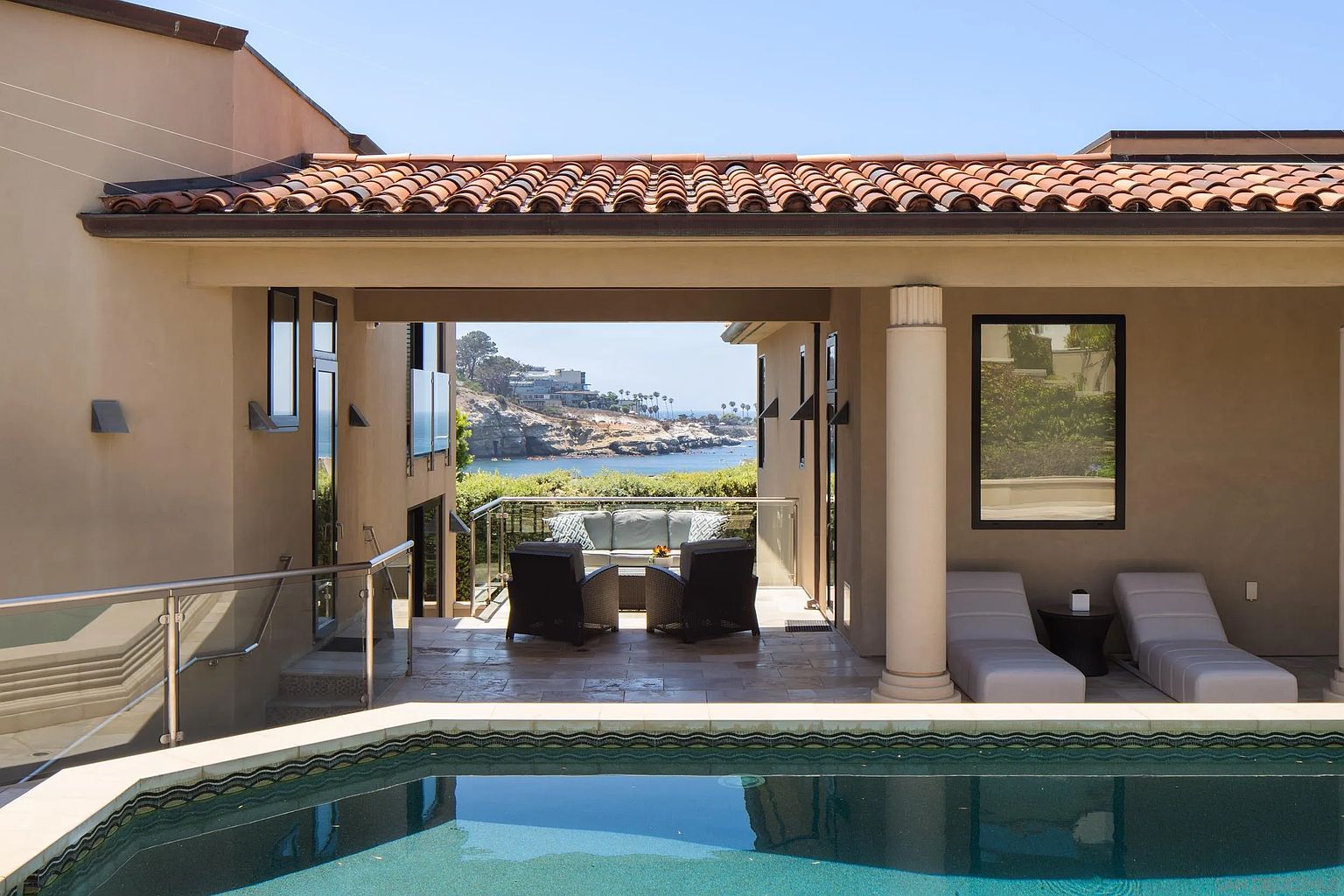 This luxurious outdoor living space features a sparkling swimming pool in the foreground, leading to a covered patio area furnished with comfortable lounge seating. The architecture showcases a Mediterranean-inspired style with terracotta roof tiles and a prominent column, framing a stunning, unobstructed view of the ocean and coastal cliffs. The scene captures a serene, high-end coastal lifestyle, perfect for relaxation and entertaining.