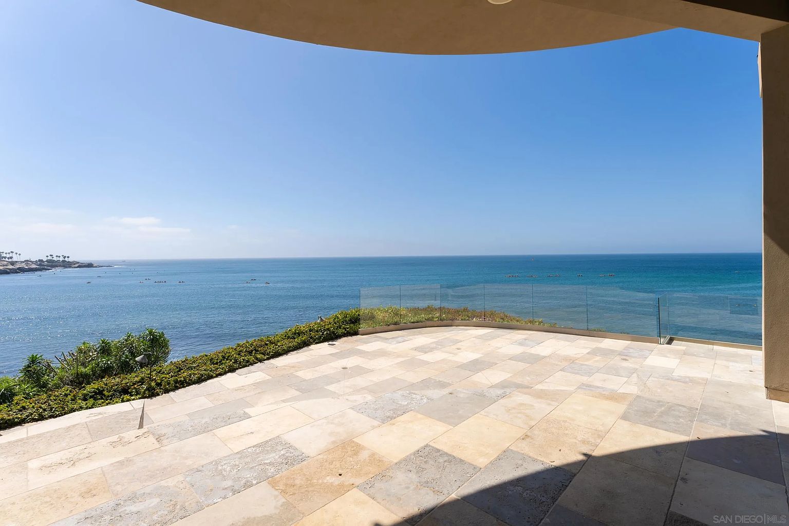 This expansive, elevated terrace offers a panoramic, unobstructed view of the ocean under a clear blue sky. The patio is paved with large, light-toned stone tiles and features a minimalist glass railing that maintains the seamless connection to the coastal horizon. Lush green landscaping borders the edge of the deck, creating a serene and luxurious outdoor living space.