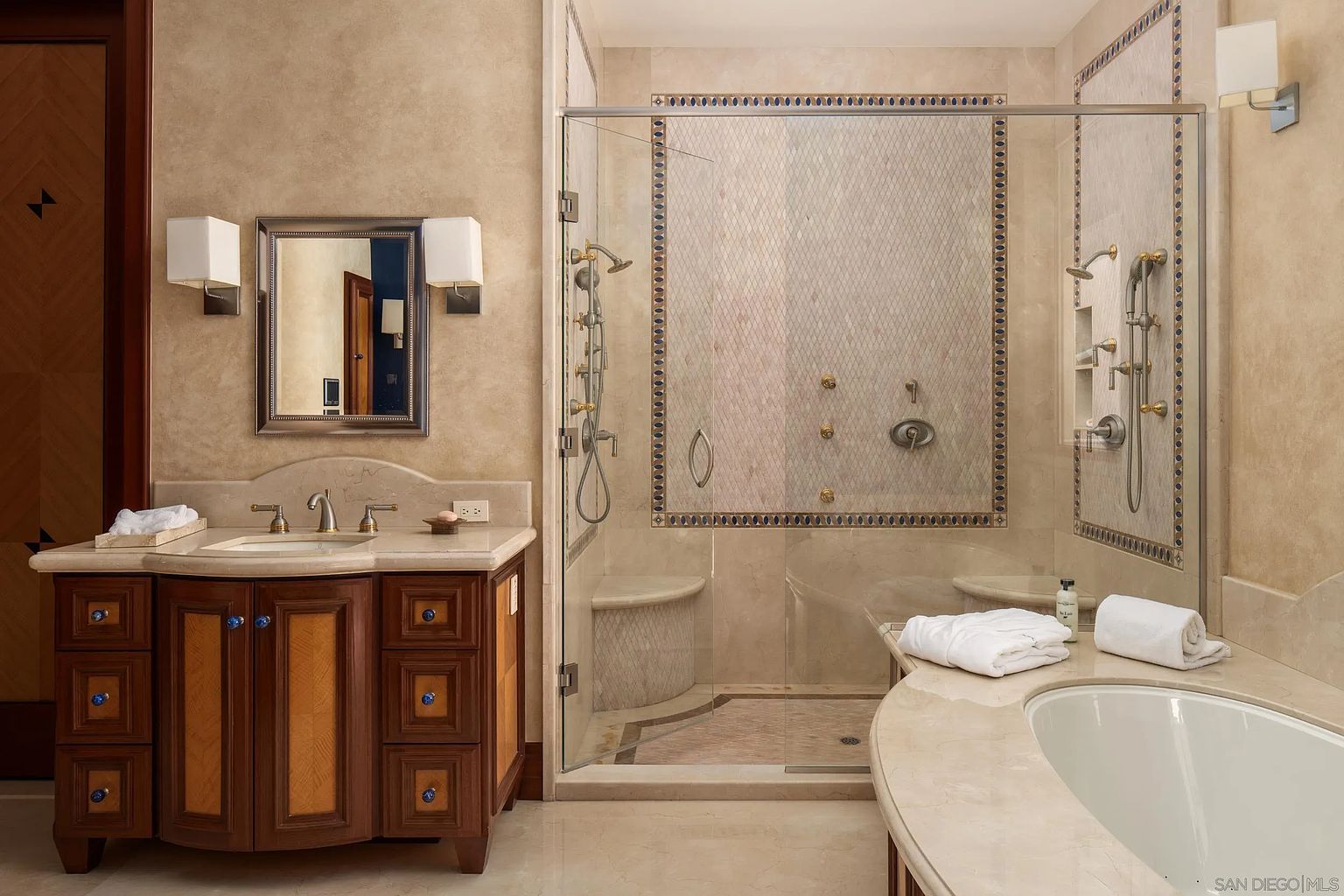 This elegant primary bathroom features a custom wood vanity with a stone countertop and a framed mirror flanked by wall sconces. A spacious walk-in shower with glass doors and intricate tile work sits adjacent to a built-in soaking tub, creating a luxurious spa-like atmosphere. The room is finished with neutral-toned walls and polished stone flooring, offering a sophisticated and cohesive design.