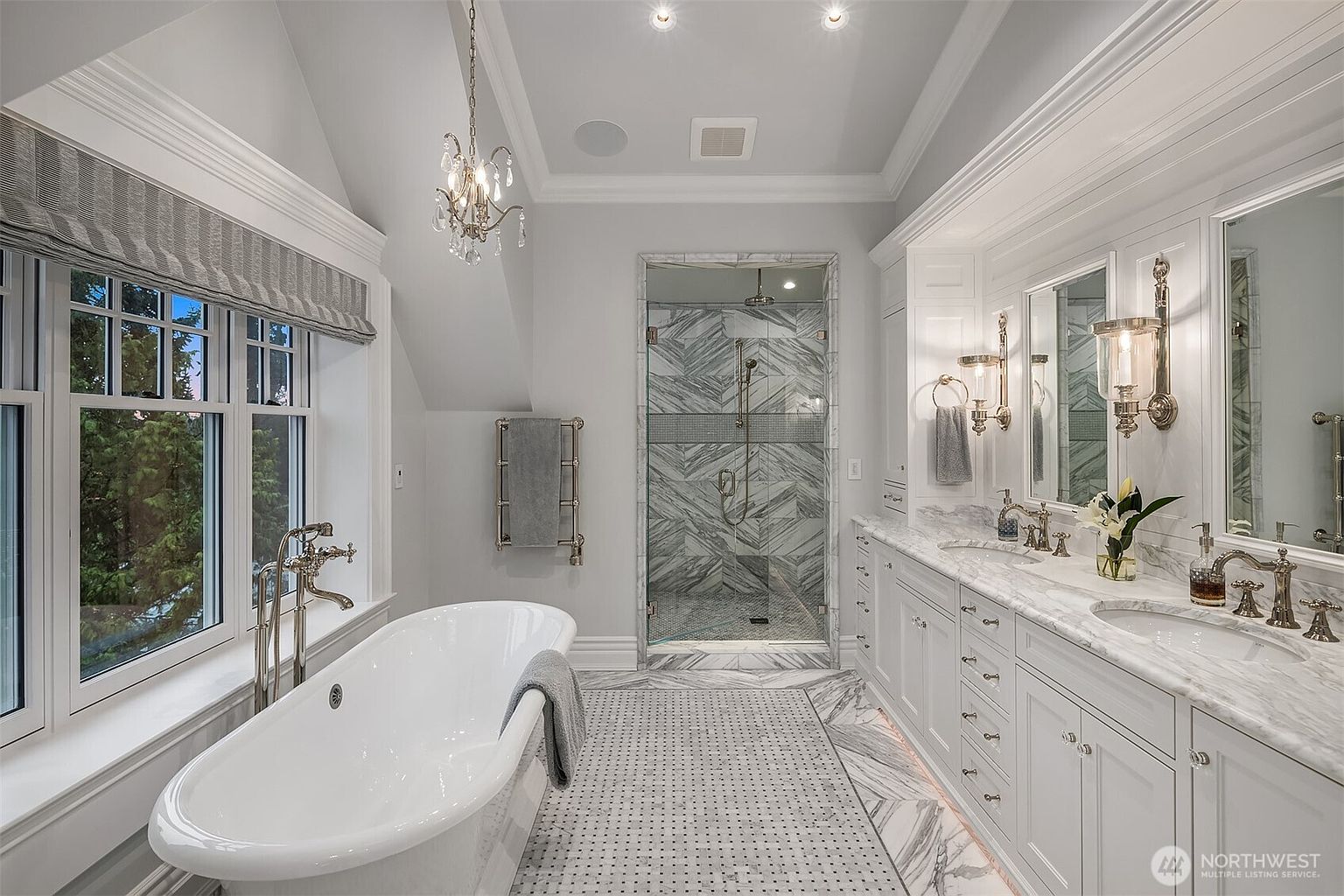 This luxurious primary bathroom features a freestanding soaking tub, a spacious marble-tiled walk-in shower, and a double vanity with elegant white cabinetry and marble countertops. The room is bathed in natural light from a large window with custom shades, complemented by sophisticated chandelier and sconce lighting. The overall aesthetic is refined and spa-like, showcasing high-end finishes and a clean, monochromatic color palette.