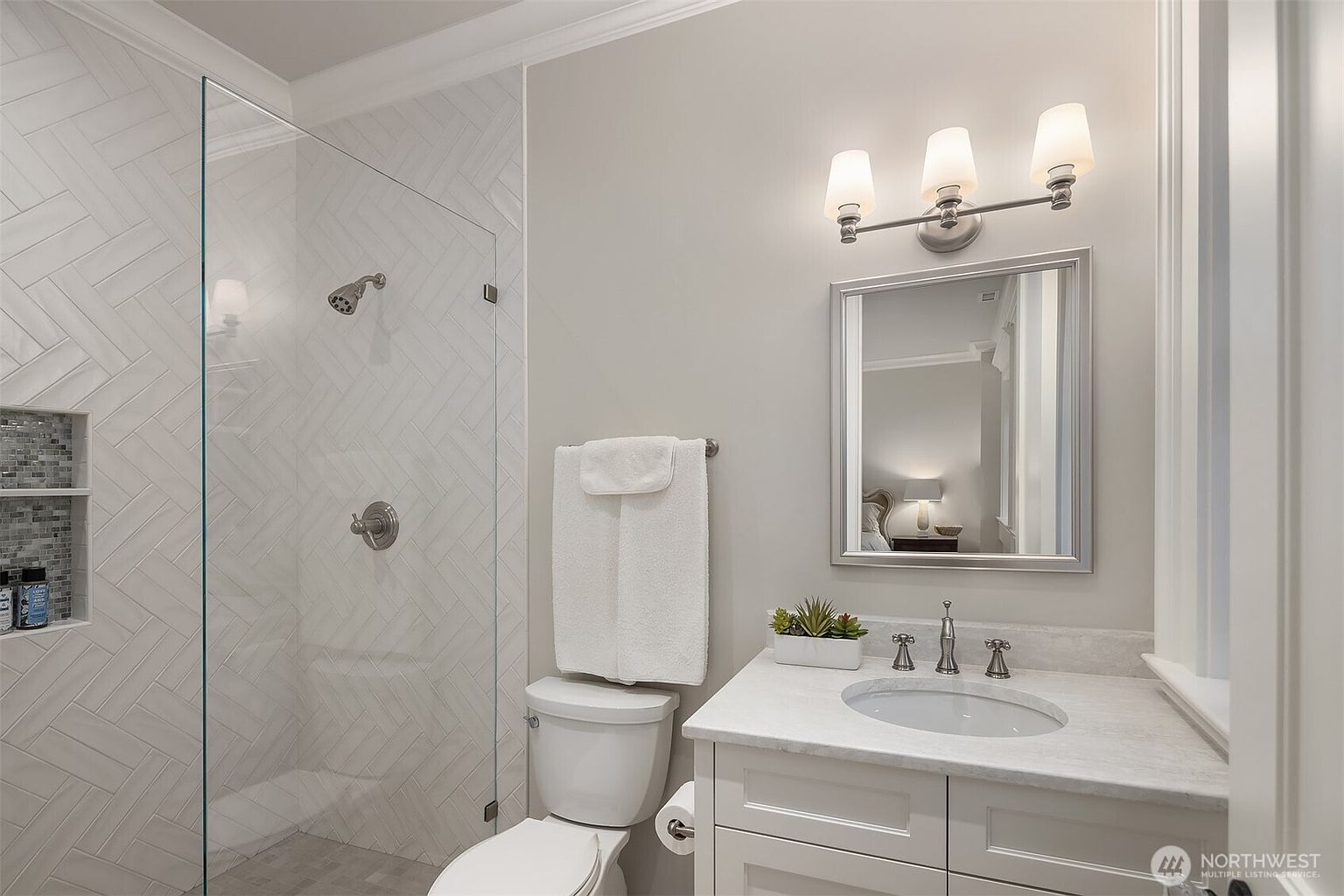 This bright and modern bathroom features a walk-in shower with a stylish herringbone tile pattern and a built-in niche. A clean white vanity with a marble countertop, a silver-framed mirror, and a three-light vanity fixture complete the space. The neutral color palette and crisp design create a clean, inviting atmosphere.