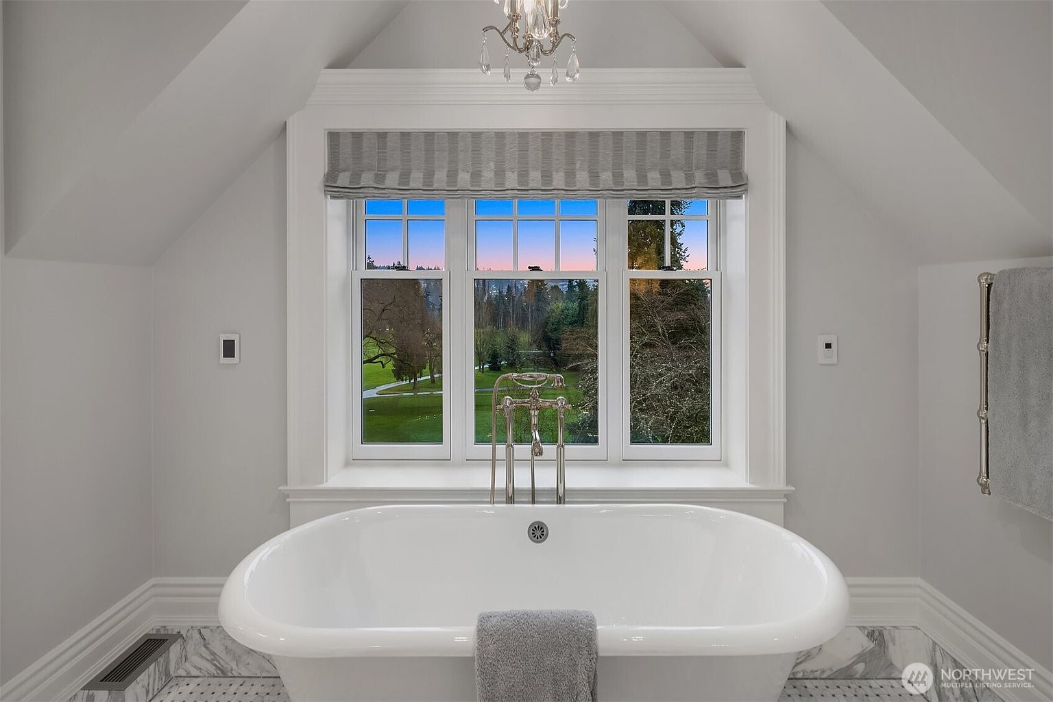 This elegant primary bathroom features a deep, freestanding soaking tub centered beneath a large, multi-pane window that offers a serene view of the lush green landscape outside. The room is finished with crisp white walls, classic crown molding, and a sophisticated marble floor, complemented by a delicate crystal chandelier overhead. A polished chrome floor-mounted tub filler and a matching towel warmer add a touch of luxury to this bright, tranquil space.