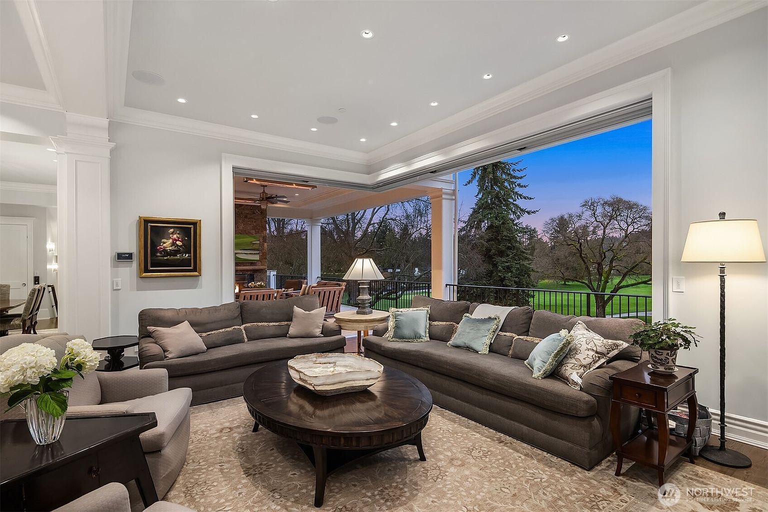 This elegant living room features a spacious, open-concept design with plush grey sofas arranged around a large, dark wood oval coffee table. The room seamlessly transitions to an expansive outdoor deck through floor-to-ceiling retractable glass walls, offering a stunning view of the lush green landscape at dusk. The sophisticated interior is accented by recessed lighting, a neutral color palette, and tasteful decor, creating a luxurious and inviting atmosphere.